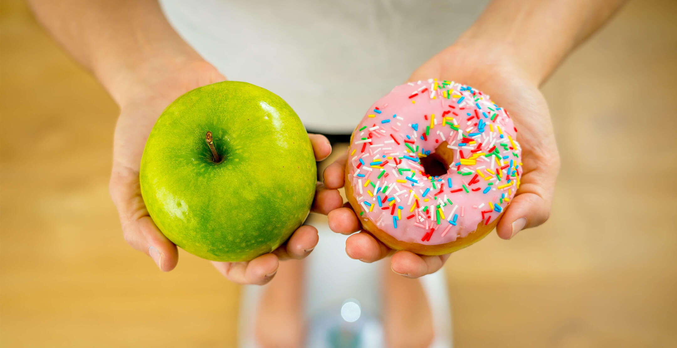 An overhead view of a person standing on a scale, holding an apple in one hand and a pink frosted donut in the other.